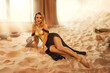 fantasy sexy woman lying posing in room full sand desert. clothes gold accessories beauty face. fashion model girl queen in black long dress golden corset armor. Sand sun divine magic light window 