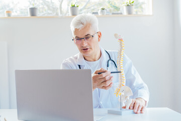 Doctor using laptop for telemedicine with patient at hospital. Osteopath makes online video call consult, pointing on human spine model. Spinal healthcare and back pain treatment. Remote appointment