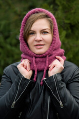  portrait of beautiful blonde smiling woman  in pink knitted handmade hood from wool posing in street near green fir tree on autumn day