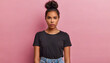serious displeased latin woman with hair bun raises eyebrows looks attentively at camera purses lips has dimple on cheek dressed in casual black t shirt and jeans isolated over pink background
