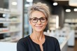 © Georgii - Portrait of a charming mature Caucasian female pharmacist wearing glasses among shelves of medicines in a pharmacy. Experienced confident professional in the workplace. Healthcare and hygiene concept.