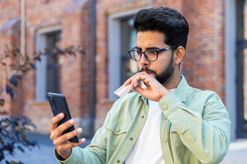 Cheated disappointed man rejected online money transfer error, businessman in casual clothes outside office building holding phone and bank credit card, internet fraud scammer.