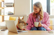 Smiling beautiful woman using laptop holding cup of coffee working in modern cafe with adorable cute dog. Successful happy freelancer sitting at workplace near lovely pet 