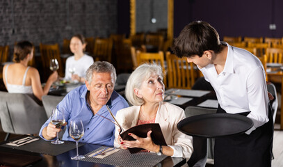 Wall Mural - Polite hospitable young waiter talking to positive elderly couple, man and woman, visiting restaurant for lunch, recommending dishes from menu card