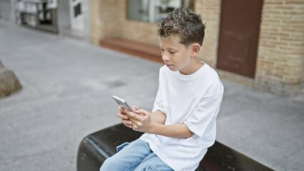 Wall Mural - Adorable blond boy totally hooked texting, seriously absorbed using phone technology, sitting relaxed on city bench, outdoor urban background