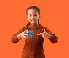 Poster - Little girl pointing at piggy bank on orange background