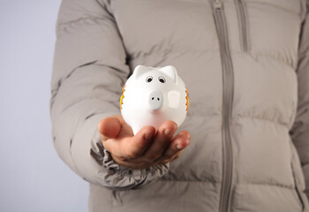 Wall Mural - Man in down jacket holds white ceramic piggy bank on gray background.