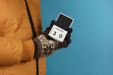 Man in yellow winter down jacket and gloves holding block calendar with date january 30 on blue background