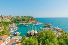 Panoramic view of Antalya, Turkey. Deep blue-green waters of the Mediterranean Sea meet a bustling harbor filled with boats of various sizes. A white lighthouse stands sentinel on a rocky outcropping