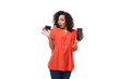 young caucasian brunette woman with curly hair dressed in an orange blouse demonstrates in surprise a plastic card and a smartphone