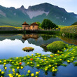 Haus am See mit vielen galben Seeblumen und Seerosen. Das Wasserhaus spiegelt sich an einem sonnigen Tag im See. Berge säumen den Hintergrund