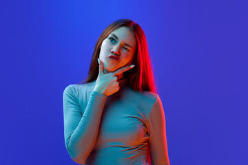 Answers and questions. Girl keeps hand on chin looking away and grimacing pensively and think or dreaming against blue background in neon light.