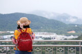 Fototapeta Dziecięca - Tourists lean against the fence to admire the beauty of the mountains and the misty morning sky. A woman wearing a yellow shirt, a red backpack and a straw hat stand beside the stainless steel fence.
