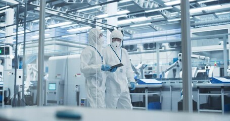 Leinwandbilder - Factory Assembly Facility: Engineers in Coveralls and Masks Walking Through Workshop Facility and Using a Tablet Computer. In Background Robotic Arms Producing Modern High Tech Electronics Components