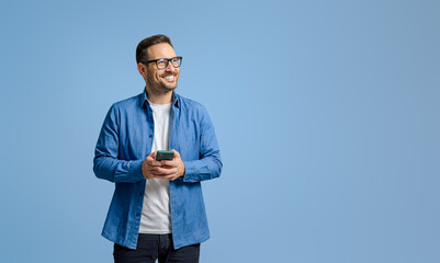 smiling businessman holding mobile phone and looking away thoughtfully against blue background