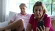 © Marco - Senior grandmother speaking with family relatives on video communication, holding smartphone device seated at home couch sofa next to her husband