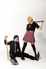 Wall Mural - man in black wig sitting  with baseball bat near woman in school uniform on white, cosplay trend