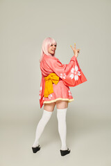 Wall Mural - young woman in blonde wig and pink kimono with white knee socks showing victory gesture on grey