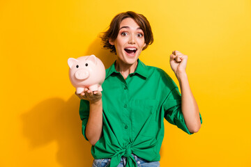 Poster - Photo of lucky excited girl dressed green shirt rising fist holding piggy cash box isolated yellow color background