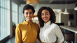 Two young female black office employees posing in modern office space, looking at camera and smiling. 