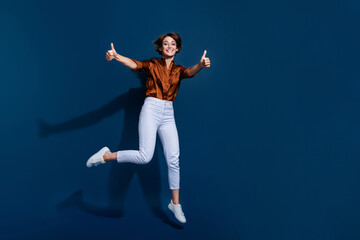 Wall Mural - Full length photo of adorable confident lady wear brown shirt jumping showing two thumbs up empty space isolated blue color background