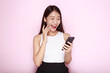 © Parichat - Portrait of a beautiful young woman in a light pink background, happy and smile, posting in stand position, Asian woman looks surprised while holding phone in hand
