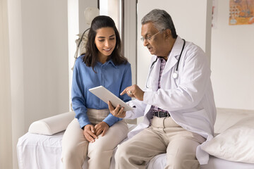 Serious mature Indian doctor explaining healthcare examination result to young patient, showing tablet computer screen to woman, sitting on medical couch, speaking, giving consultation