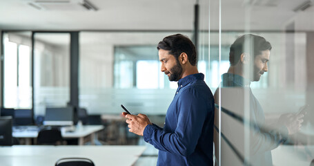 Busy Indian business man holding mobile phone device looking at cellphone standing at work. Professional businessman using finance banking apps on smartphone working on cell phone in office. Side view