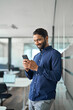 © insta_photos - Busy Indian businessman using mobile cell phone working standing in office. Smiling professional business man making banking payment in app on smartphone working on cellphone at work. Vertical photo.