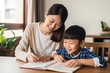 © Stavros - Mom helps son do homework sitting at table writing in notebook right answer. Boy asks mother to help with homework at table with laptop. Mom in cozy modern apartment helps her son with lessons