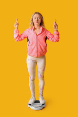 Poster - Beautiful sporty woman with crossed fingers and scales on yellow background. Weight loss concept