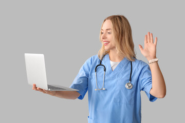 Wall Mural - Beautiful female doctor with laptop waving hand on grey background