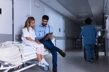 Diverse male doctor talking with girl using tablet on bed in hospital corridor