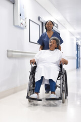 Wall Mural - African american female doctor walking with senior female patient in wheelchair in hospital corridor