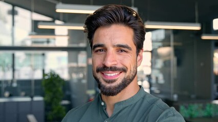 Wall Mural - Happy young Latin business man looking at camera in office, headshot portrait. Smiling bearded businessman, male entrepreneur, professional employee looking at camera standing at work.