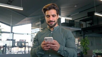 Wall Mural - Busy Latin business man executive using smartphone standing in office. Smiling handsome young businessman employee holding cell phone working on mobile texting message standing in office.