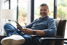 Portrait Of Happy Senior Biracial Man Sitting In Armchair And Reading Book At Home