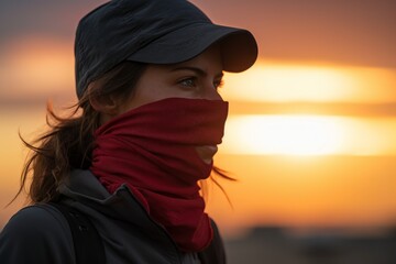 Wall Mural - Portrait of a content woman in her 30s wearing a protective neck gaiter against a vibrant sunset horizon. AI Generation