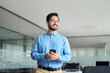 © insta_photos - Happy young latin business man holding smartphone standing in office. Smiling hispanic businessman entrepreneur or manager using finances management app on cell phone technology looking away at work.