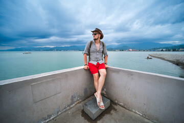 A man in red swimming shorts, with a backpack, a hat and sunglasses, sits on the waterfront and enjoys the view. Black Sea Batumi, Georgia