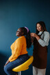 © gstockstudio - Confident African hairdresser braiding hair to a female customer on blue background