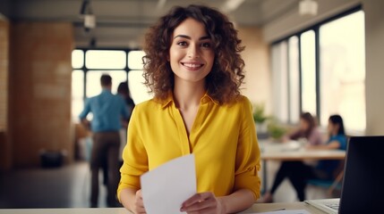 Young beautiful woman in yellow shirt studying on desk looking to camera in modern office