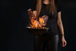Female with tattooed hands, in leggings and t-shirt. Holding wok pan with fire and frying two beef steaks against black background. Close up