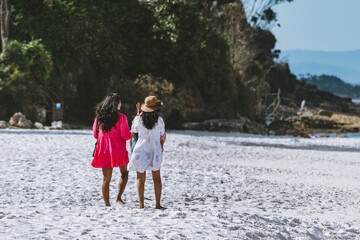 Wall Mural - two adult women wearing dresses and hats to cover themselves from the sun walking along the white sand beach towards a set of green trees in the background