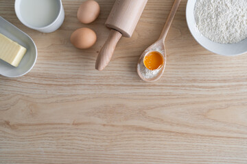 Baking flat lay composition with copy space on a wooden kitchen table or a kneading board. Rolling pin, wood spoon utensils and fresh dough ingredients for a homemade cake - flour, butter, eggs, milk.