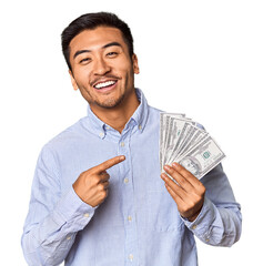 Young Chinese man holding dollar bills in studio smiling and pointing aside, showing something at blank space.