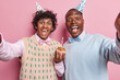 Horizontal shot of happy positive men hold cupcake with candle feel positive dressed in festive clothing and cone hats laugh happily isolated over pink background. People and celebration concept