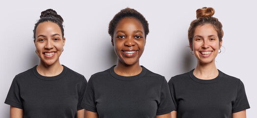Wall Mural - Horizontal shot of three cheerful young women smile gladfully look directly at camera express positive emotions feel very glad dressed in casual black t shirts isolated over white background