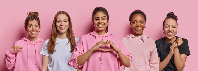 Wall Mural - Positive female friends smile toothily express happy emotions make heart gesture express love dressed in casual hoodies and t shirts being in good mood isolated over pink background. Collage image