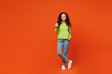 Full body young woman of African American ethnicity she wear hoody casual clothes hold takeaway delivery craft paper brown cup coffee to go isolated on plain red orange background. Lifestyle concept.